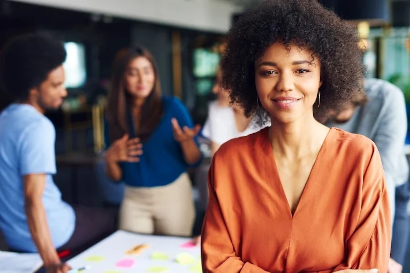 A imagem mostra uma mulher em primeiro plano, usando uma blusa laranja vibrante, com uma expressão serena e confiante. Ela tem cabelos cacheados e está com os braços cruzados, transmitindo uma sensação de liderança e autoconfiança. Ao fundo, levemente desfocados, estão outras três pessoas em uma interação, discutindo em torno de uma mesa cheia de post-its coloridos. O ambiente parece ser de trabalho colaborativo, com um ar informal e criativo. A mulher em laranja se destaca como uma figura central e segura, simbolizando a liderança feminina no contexto de equipe e inovação.