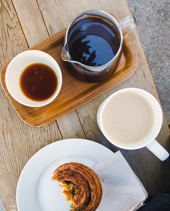A tray with 2 cups of coffee, a mug, and a half eaten pastry sitting on a bench.