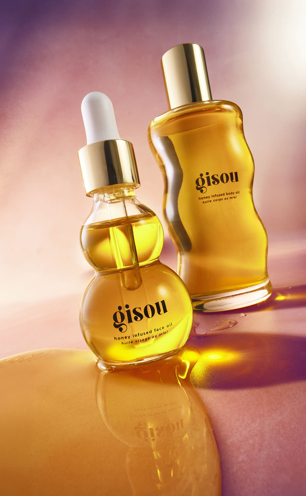 Two bottles of golden oil are displayed against a soft gradient background, highlighting their luxurious appearance.