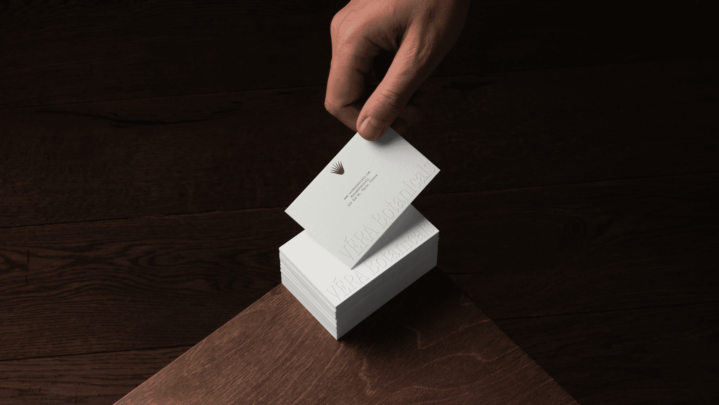 A stack of business cards sitting on the corner of a table. A hand is reaching out to grab the card on the top of the stack. The cards feature the VÉRA Botanicals logo embossed on the bottom and the company details printed in the center of the card.