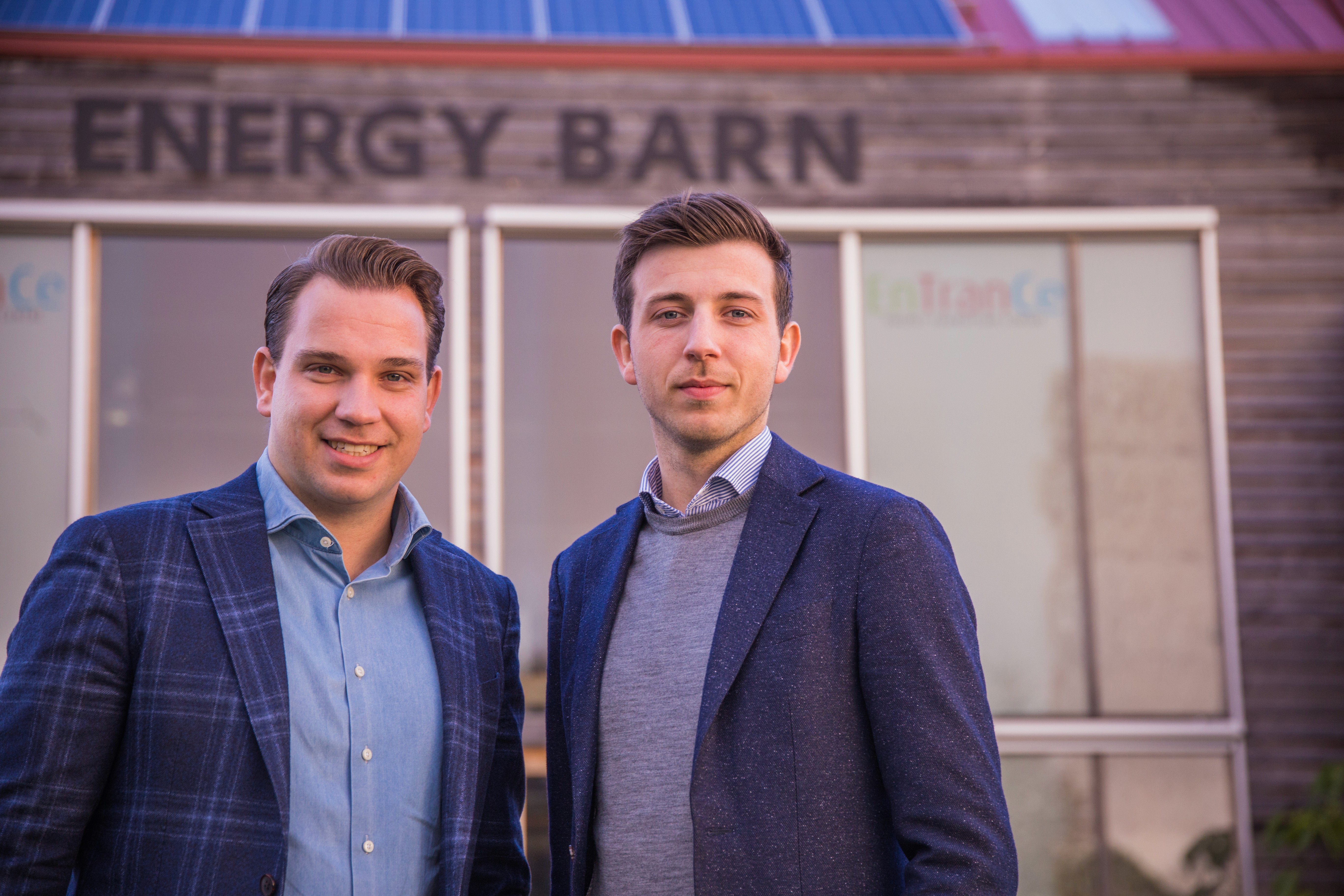 A young person with curly hair smiles at the camera while sitting in front of a laptop against a dark background.