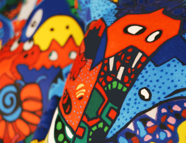 A close-up view of a vibrant textile with a colorful, hand-drawn design. The fabric features bold shapes and patterns in bright reds, oranges, yellows, blues, and greens, with white accents. The texture appears slightly raised or embossed, giving it a three-dimensional quality. The design includes abstract elements and possibly stylized floral or geometric shapes, creating a dynamic and playful visual effect.