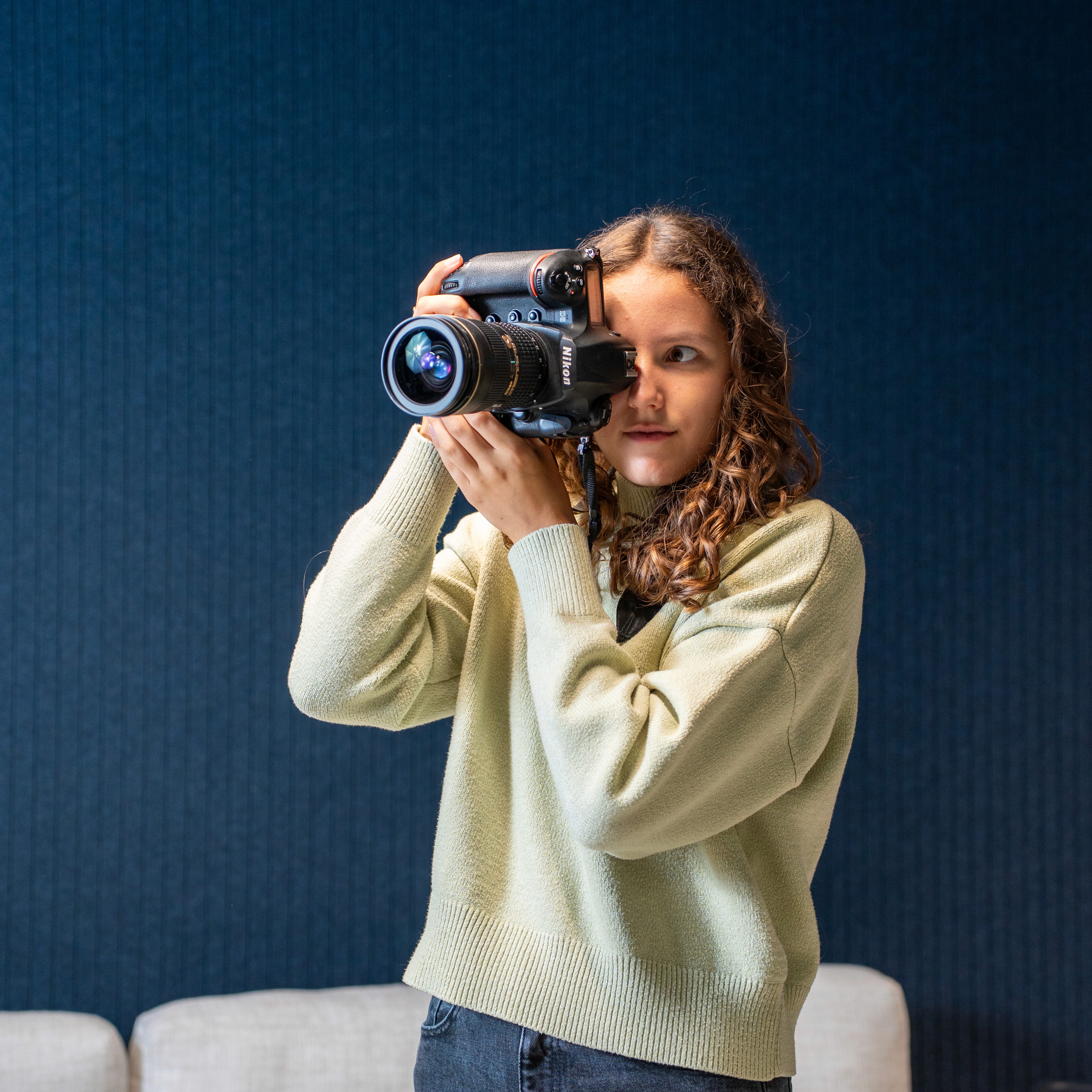 A young person with curly hair smiles at the camera while sitting in front of a laptop against a dark background.