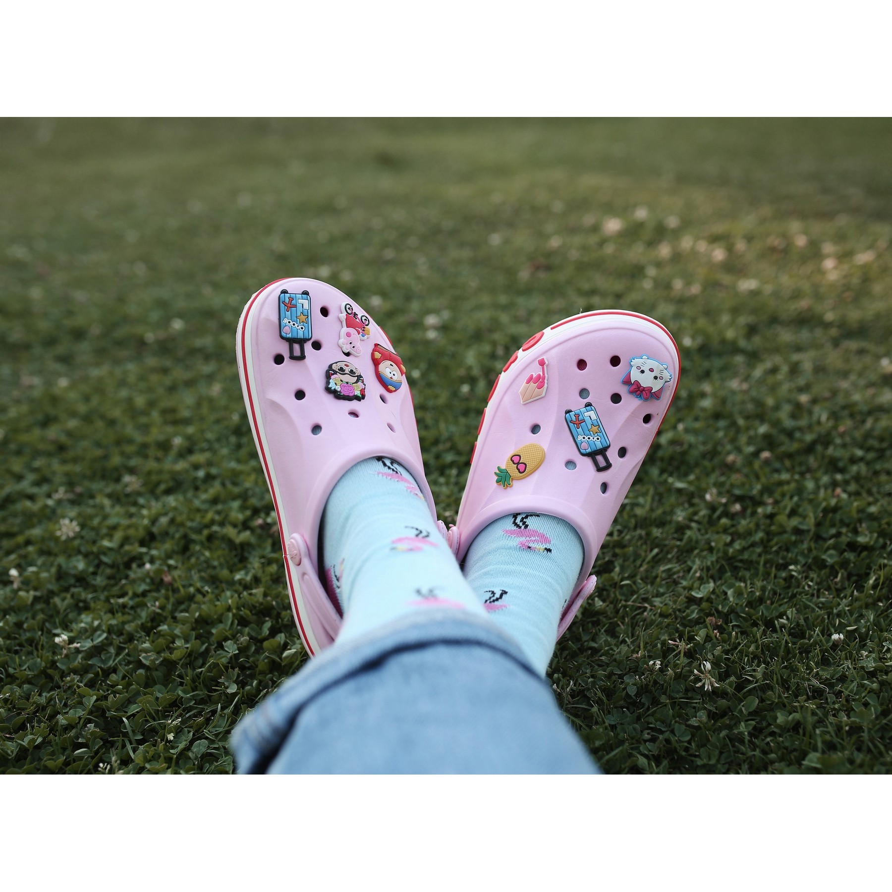 Two legs crossed wearing crocs with custom shoe charms on grass.
