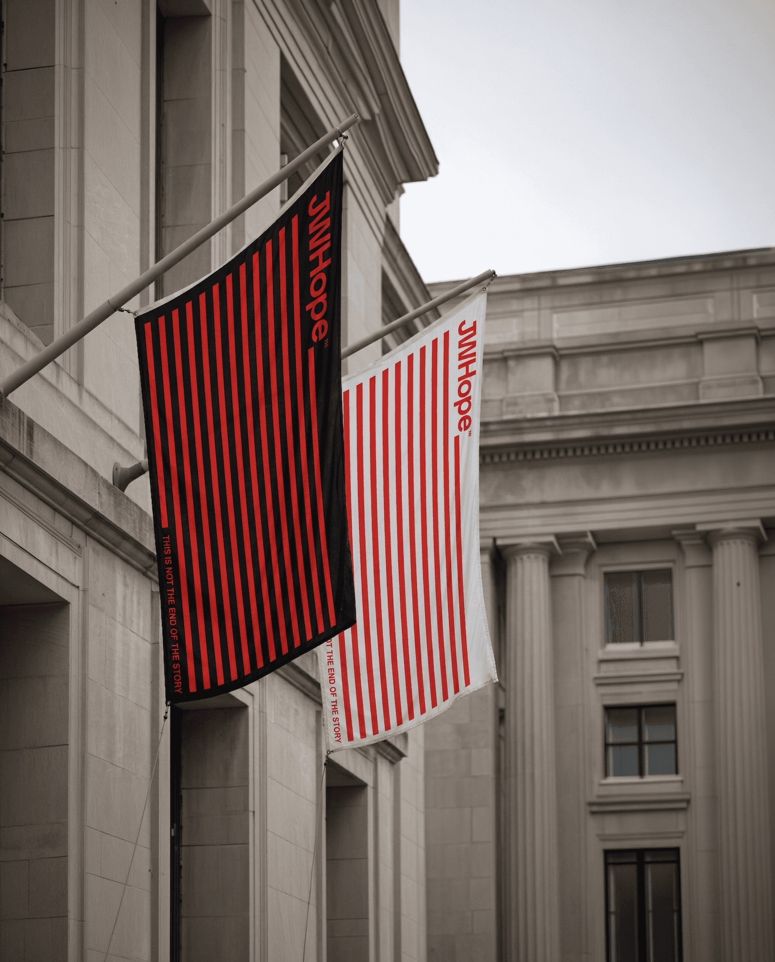 Two vertically striped flags, one red on black and the other red on white, hang from a grand neoclassical building with impressive columns and windows.