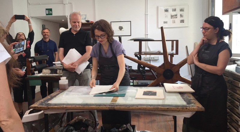 A photo of a group of people looking at a woman how she prepares the paper imprints on a table.