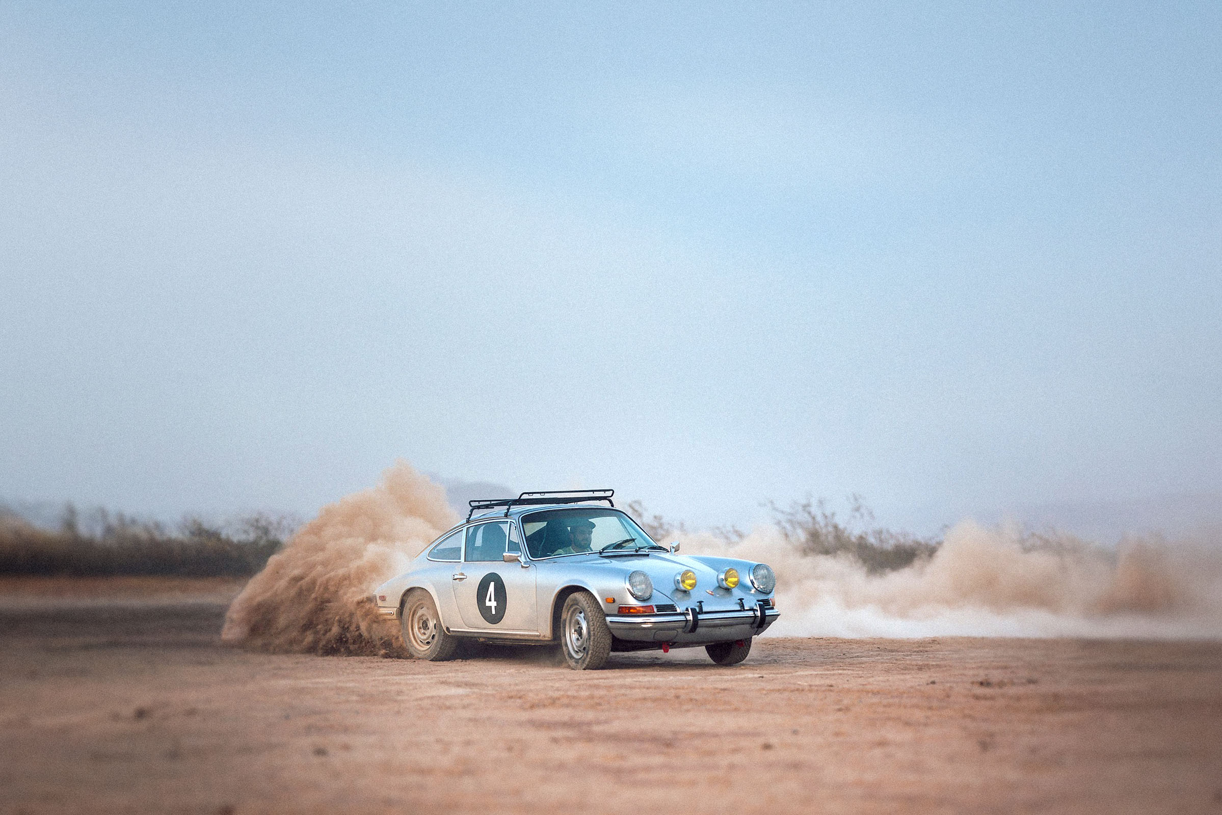 A vintage silver Porsche rally car with the number 4 on its side kicks up a cloud of dust as it drifts on a dirt track in a desert landscape.