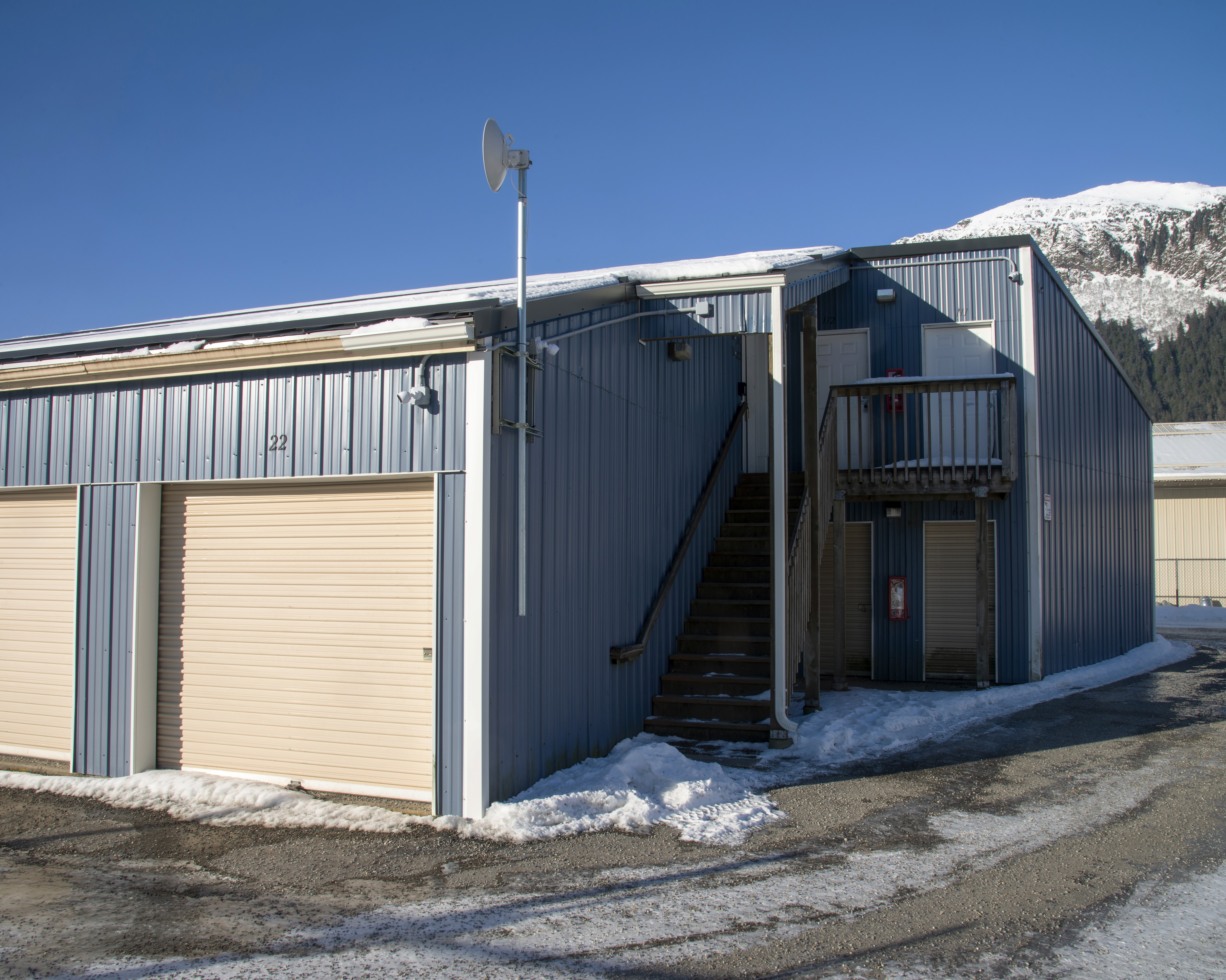 Exterior corner view of a grey metal self-storage building with a beige roll-up door, an exterior staircase with metal railings leading to a second-floor office or manager unit, a satellite dish mounted on a pole, and snow patches on the ground against a clear blue sky and mountain backdrop.