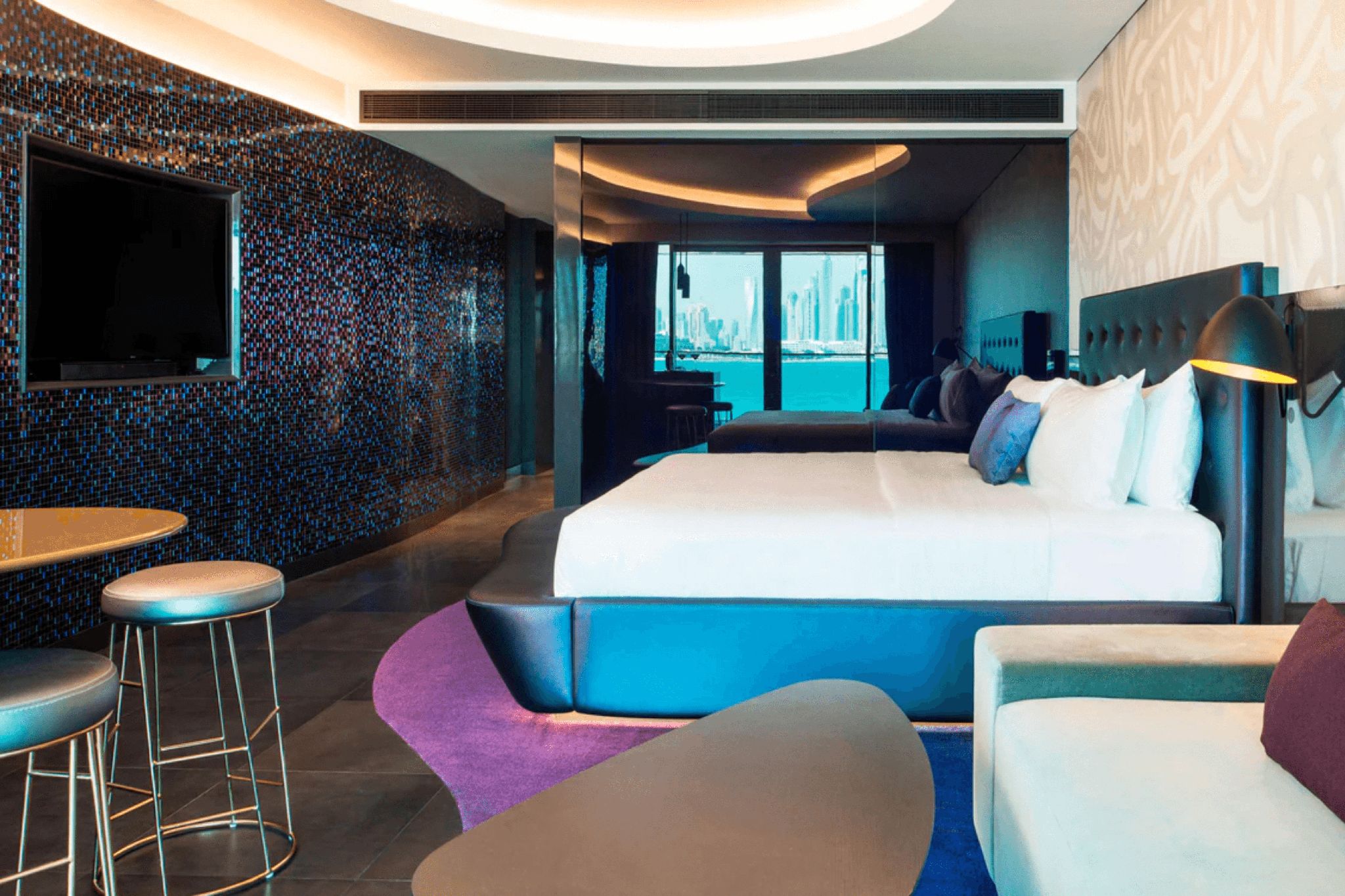 A room in W Dubai at The Palm with a colorful wall design, bar stools and table, and a large bed.