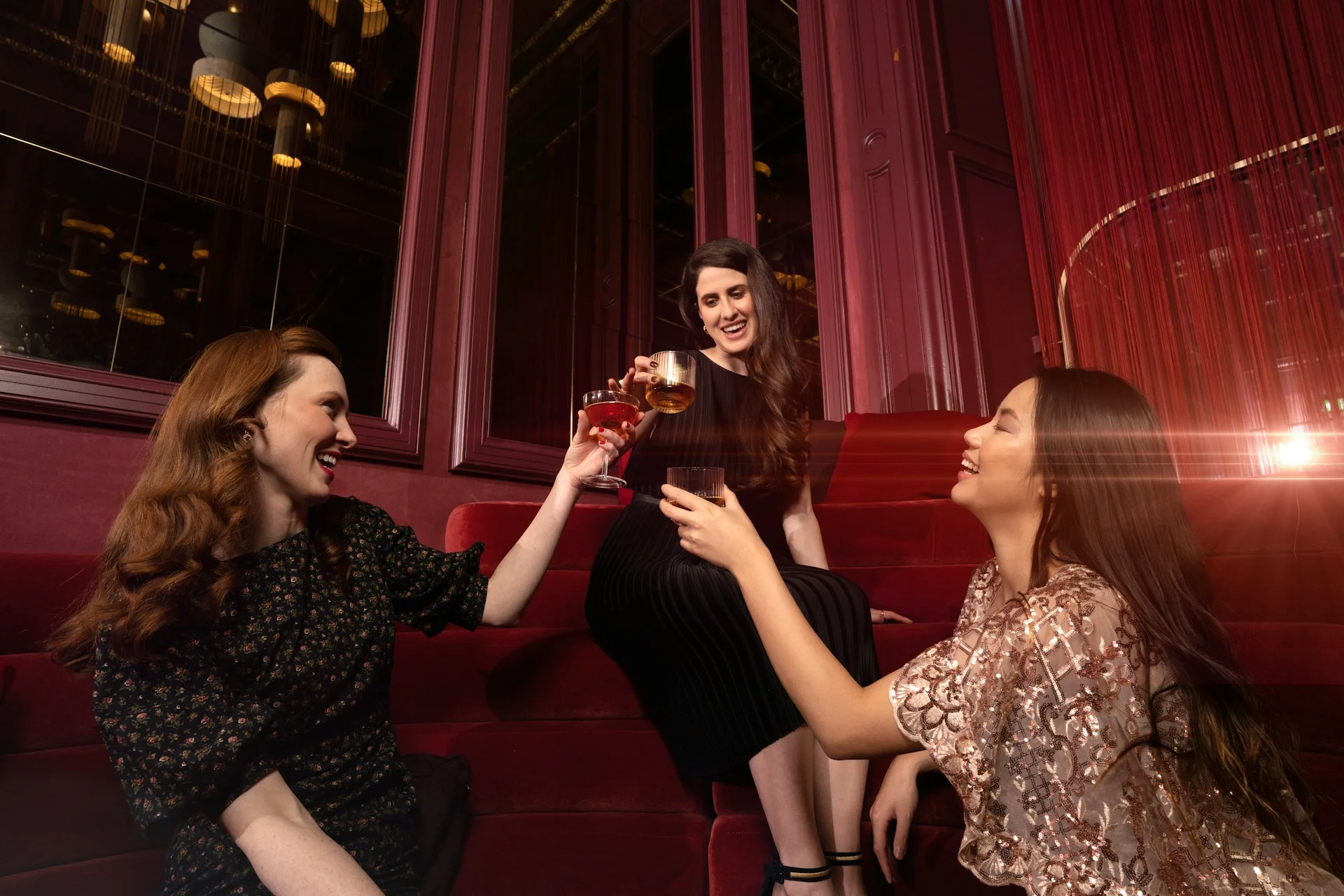 Three women enjoying cocktails together in a stylish red lounge interior with evening lighting.