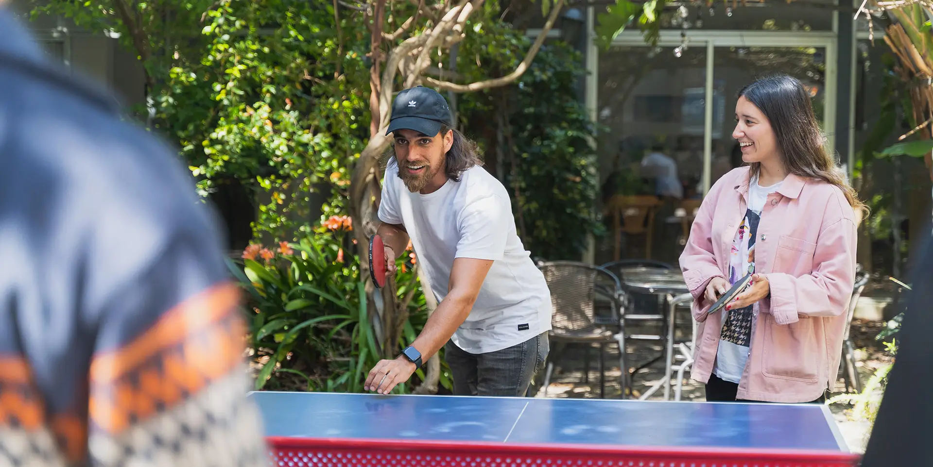Dos personas jugando tenis de mesa mientras otra los observa. De fondo se ven árboles