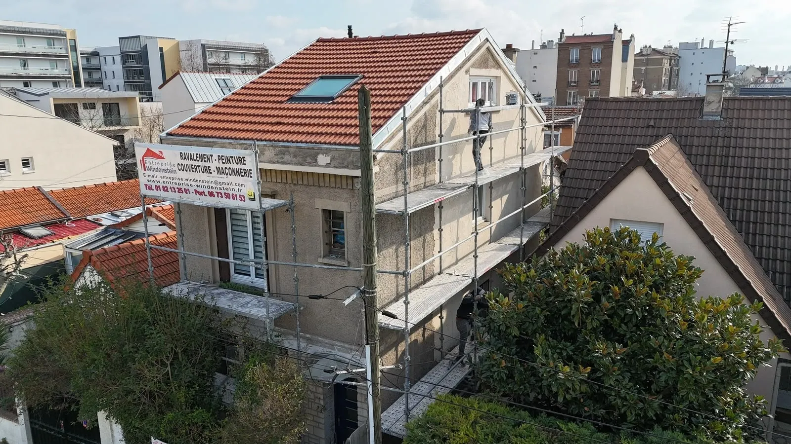 Maison à la façade beige avec un échafaudage, un panneau Entreprise Windenstein et une toiture en tuiles rouges