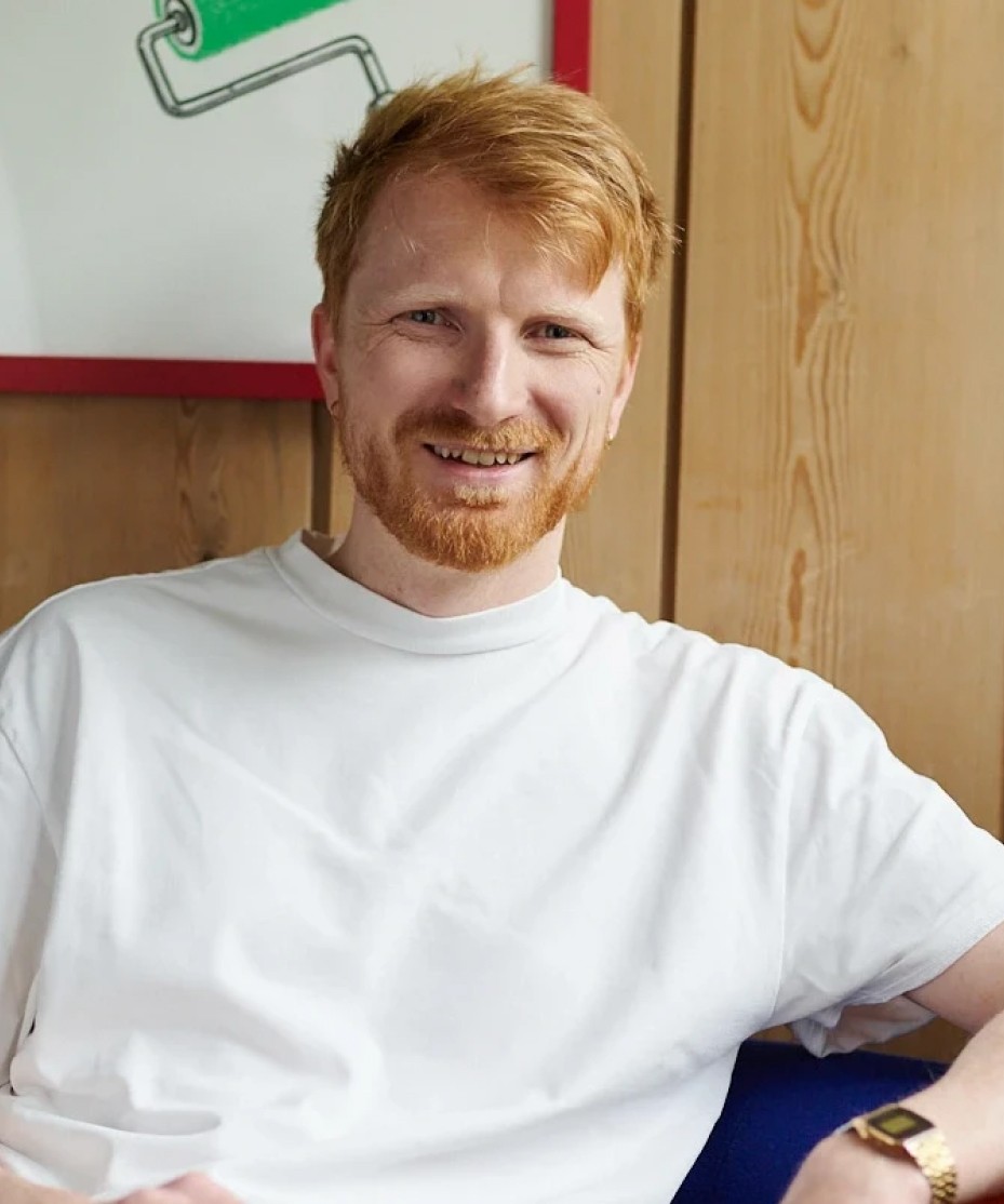 Portrait of Anders Bakken, sitting in front of a wooden wall, smiling invitingly.