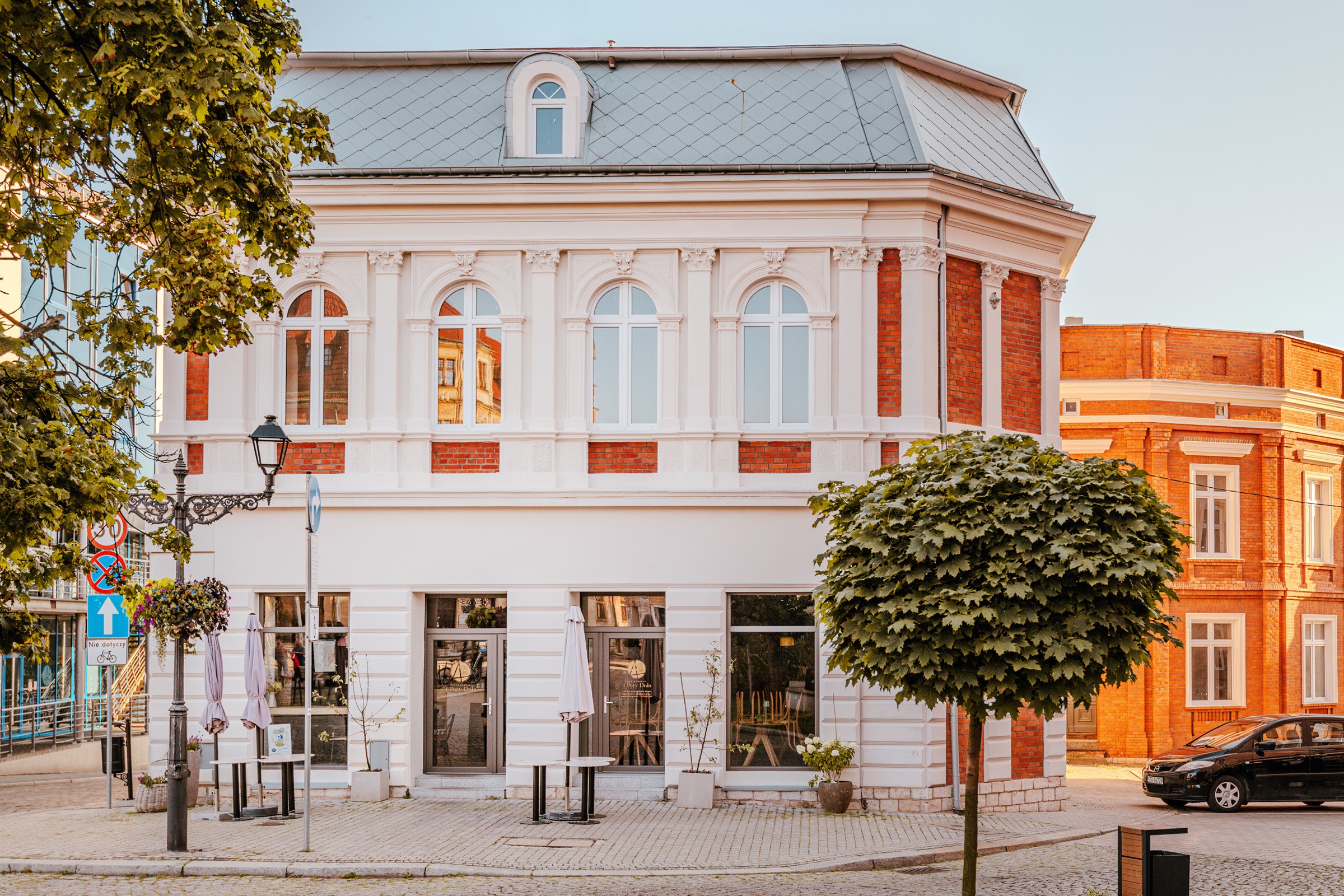 Piękny budynek w stylu klasycystycznym z czerwonymi cegłami. Na rogu ulicy z białymi elementami. Stoliki i parasole na zewnątrz.
