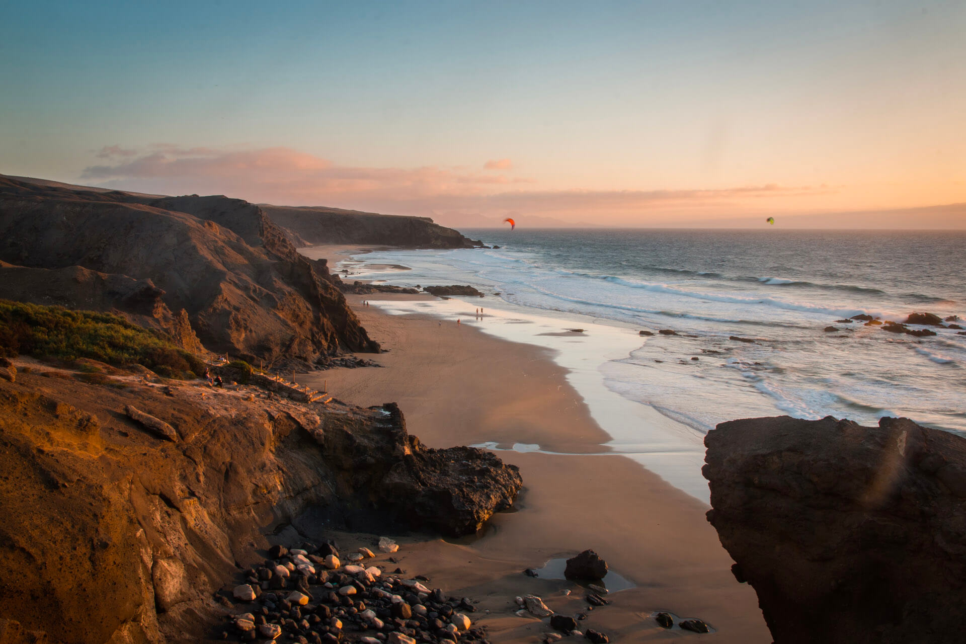 Beach with cliffs and kitesurfers at sunset.