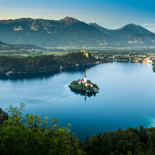 נוף ציורי של אגם שקט עם אי קטן שבו כנסייה, מוקף בהרים תחת שמיים כחולים ובהירים.