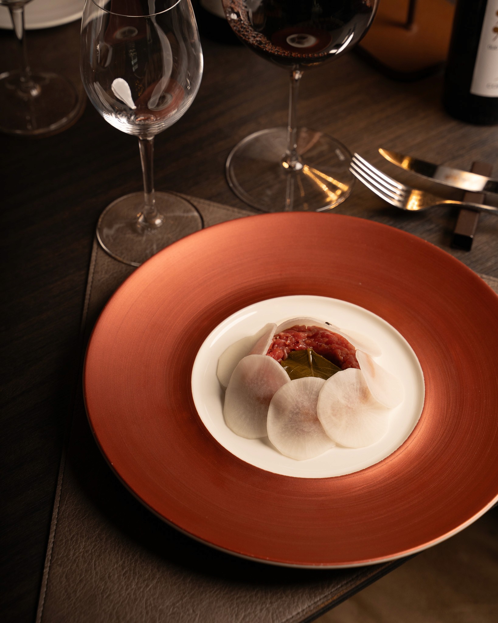 „Elegantes Gericht mit feinem Rindertatar, serviert unter dünn geschnittenen Radieschenscheiben auf einem weißen Teller mit rotbraunem Rand, begleitet von Rotwein und edlem Besteck.
