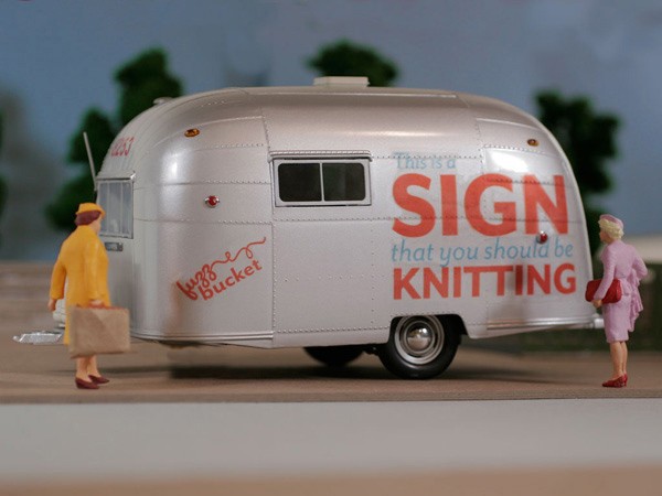 A white model Airstream trailer is parked on a surface. The side of the trailer features the text "This is a SIGN that you should be KNITTING" in red and blue letters. Two miniature figures stand near the trailer: one in a yellow coat holding a paper bag near the front, and another in a pink dress holding a red purse near the rear. The scene is set against a light blue backdrop with green foliage visible in the background.