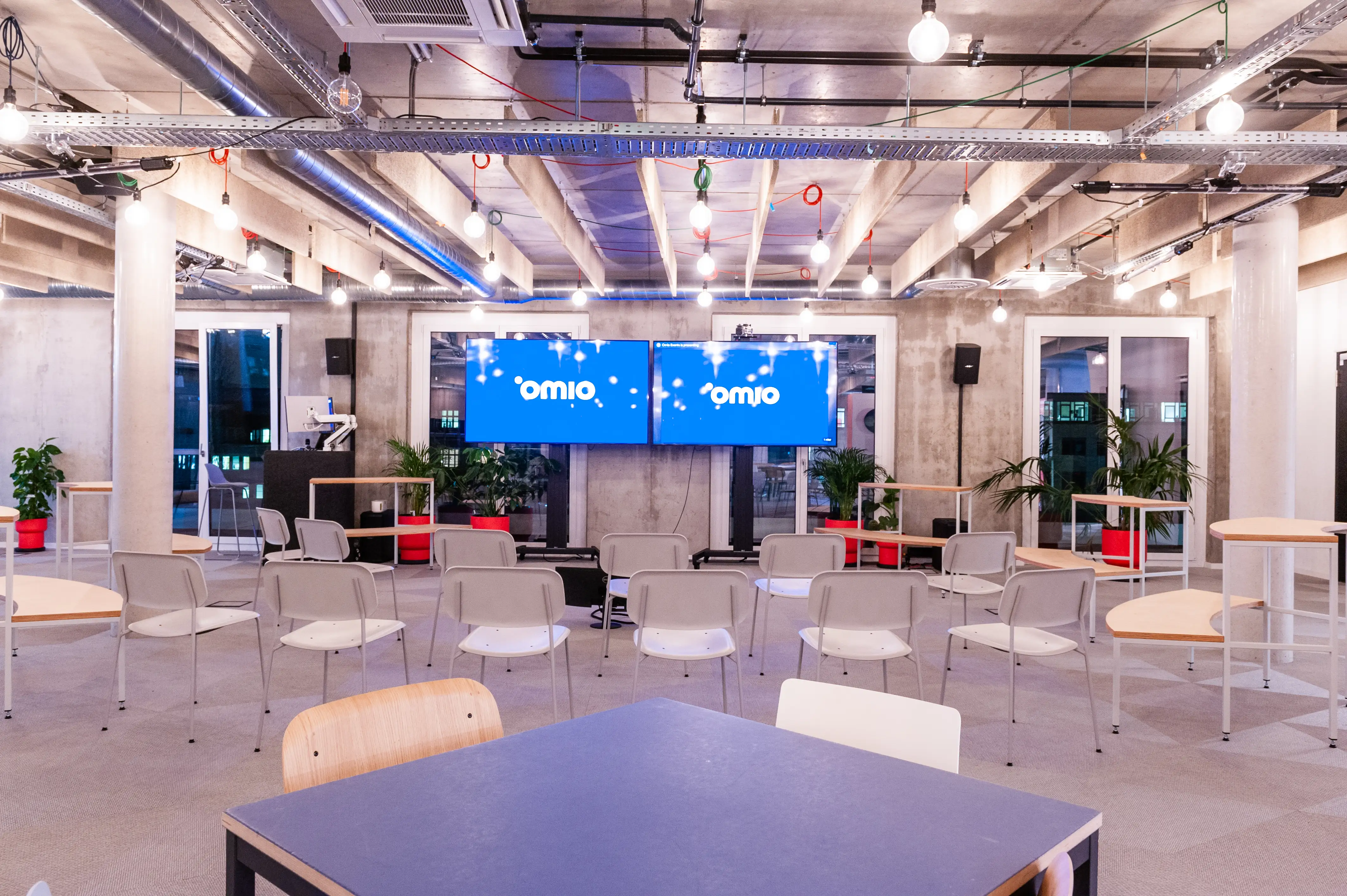 Event space at OMIO with rows of light grey chairs facing two screens showing the Omio logo, speakers on stands, red and white side tables and exposed concrete ceiling with cable trays.