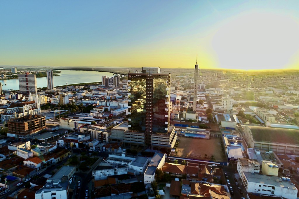 Vista aérea do Trade Center Dom Campelo em Petrolina-PE, edifício empresarial moderno com fachada de vidro espelhado escuro e detalhes brancos nas laterais, destacando-se na paisagem urbana com o Rio São Francisco e a ponte ao fundo, cercado por construções baixas do centro da cidade.