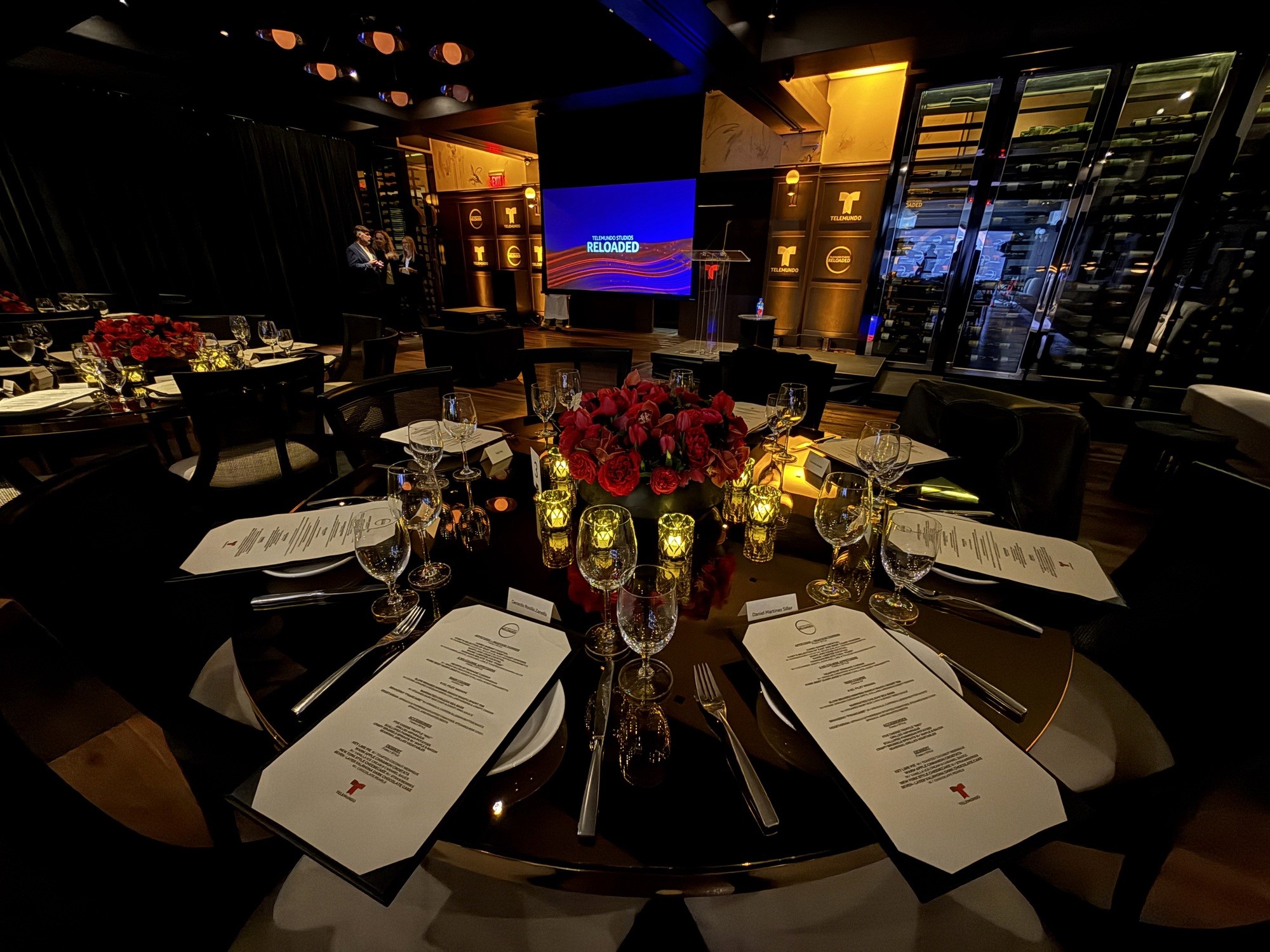 Luxury private dinner setup with glossy black tables, red floral centerpieces, and presentation screen.