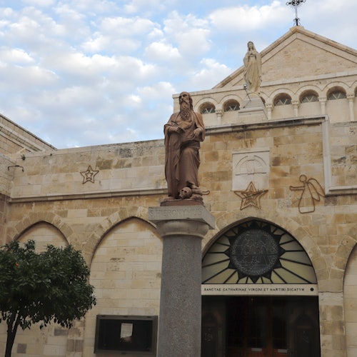 A statue on a pedestal in front of a stone building with religious symbols and a tree on the left side.