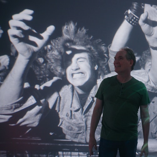 A man in a green shirt stands in front of a large vintage photo of enthusiastic concert-goers with raised hands.