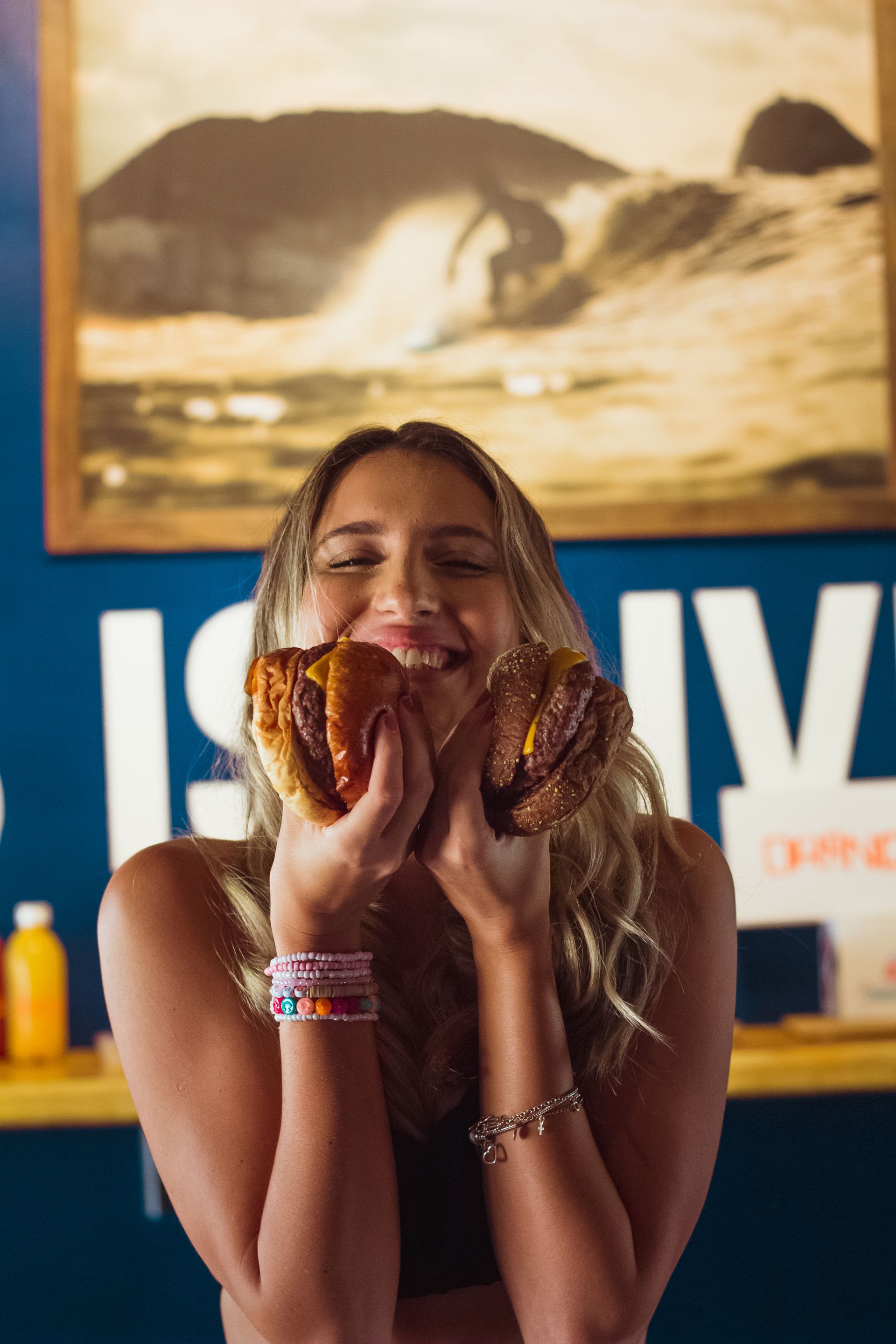 A cheerful young blonde woman holding two cheeseburgers up near her face and smiling broadly, standing indoors in front of a blue wall and a framed surfing picture.