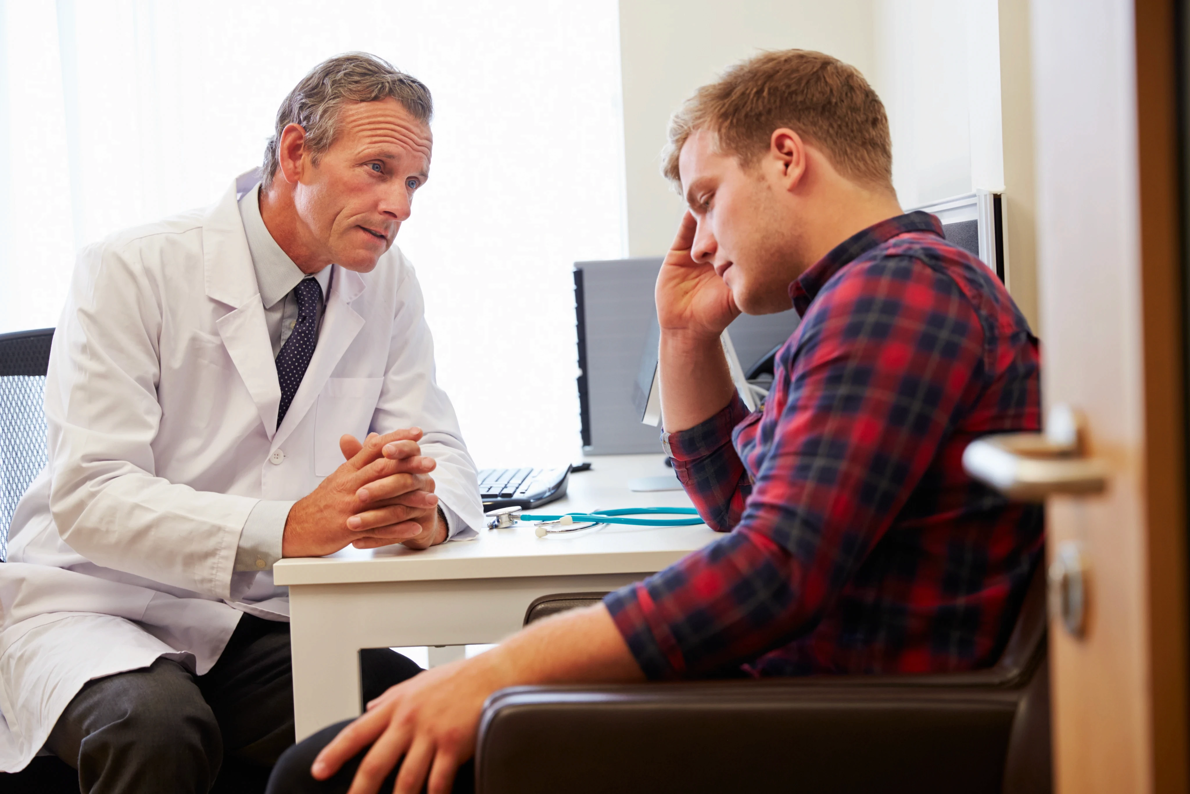 Doctor in white coat consulting a male patient in a plaid shirt in a medical office with a stethoscope on the desk.