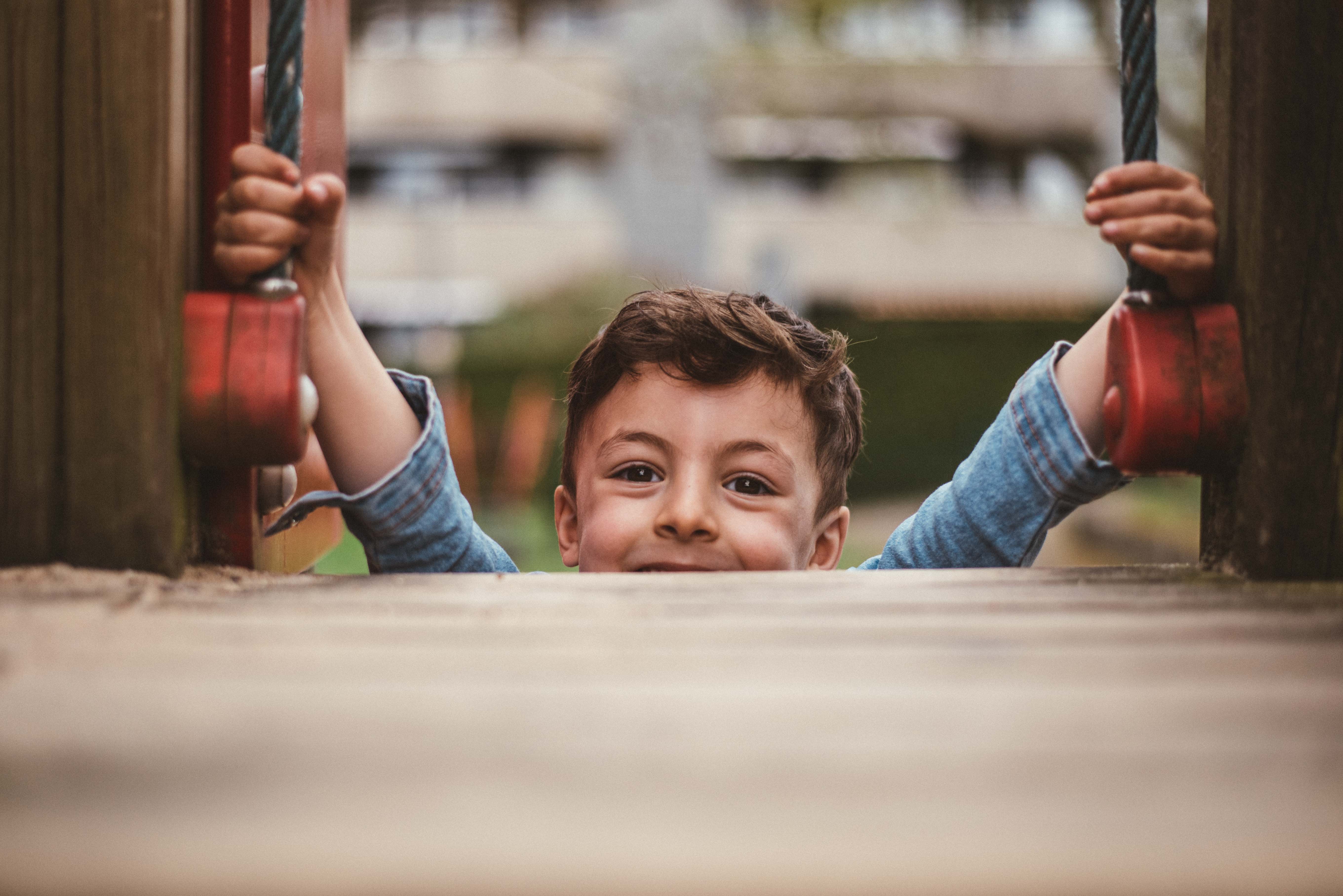 Authentische Kindergartenfotografie Gevelsberg: Ein Mädchen in einem rosafarbenen Pullover lacht glücklich beim Klettern auf einem Holzgerüst. Professionelles Outdoor-Porträt mit weichem Bokeh von PK photography 4 Kids NRW.