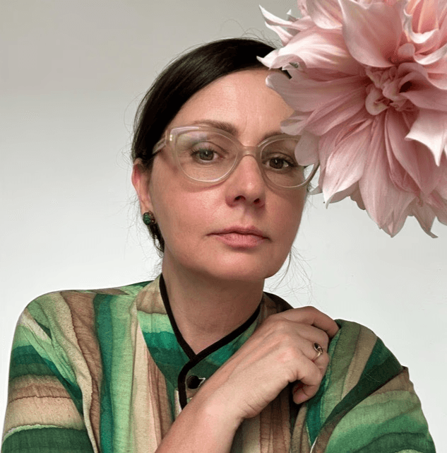 Retrato de Irina Khatsernova con gafas transparentes y blusa verde, acompañada por una flor rosada grande en primer plano.