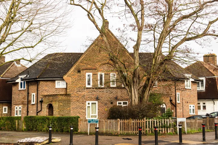 A smiling man with curly hair is sitting at a desk, looking at the camera, with a laptop and a lamp in the background.