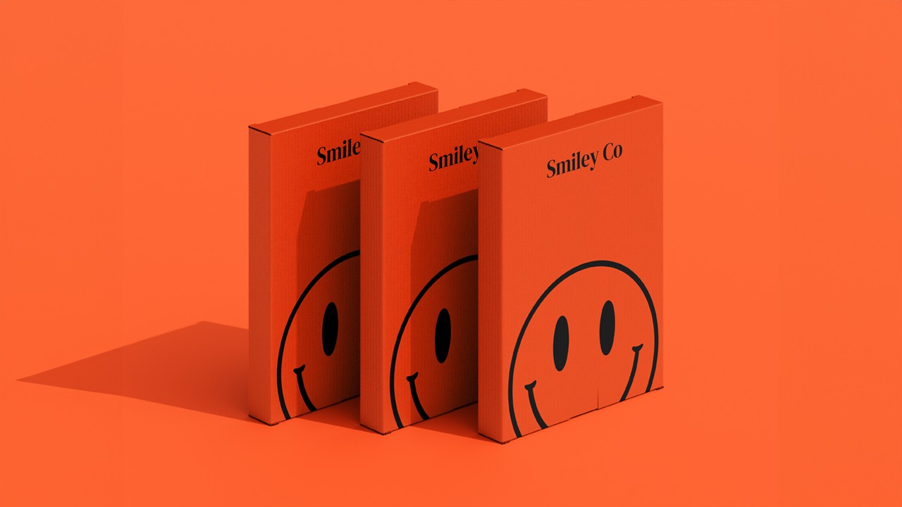 Three slim, upright orange packaging boxes side by side, each showing a partial black smiley face and “Smiley Co” text on the top, with a matching orange background.