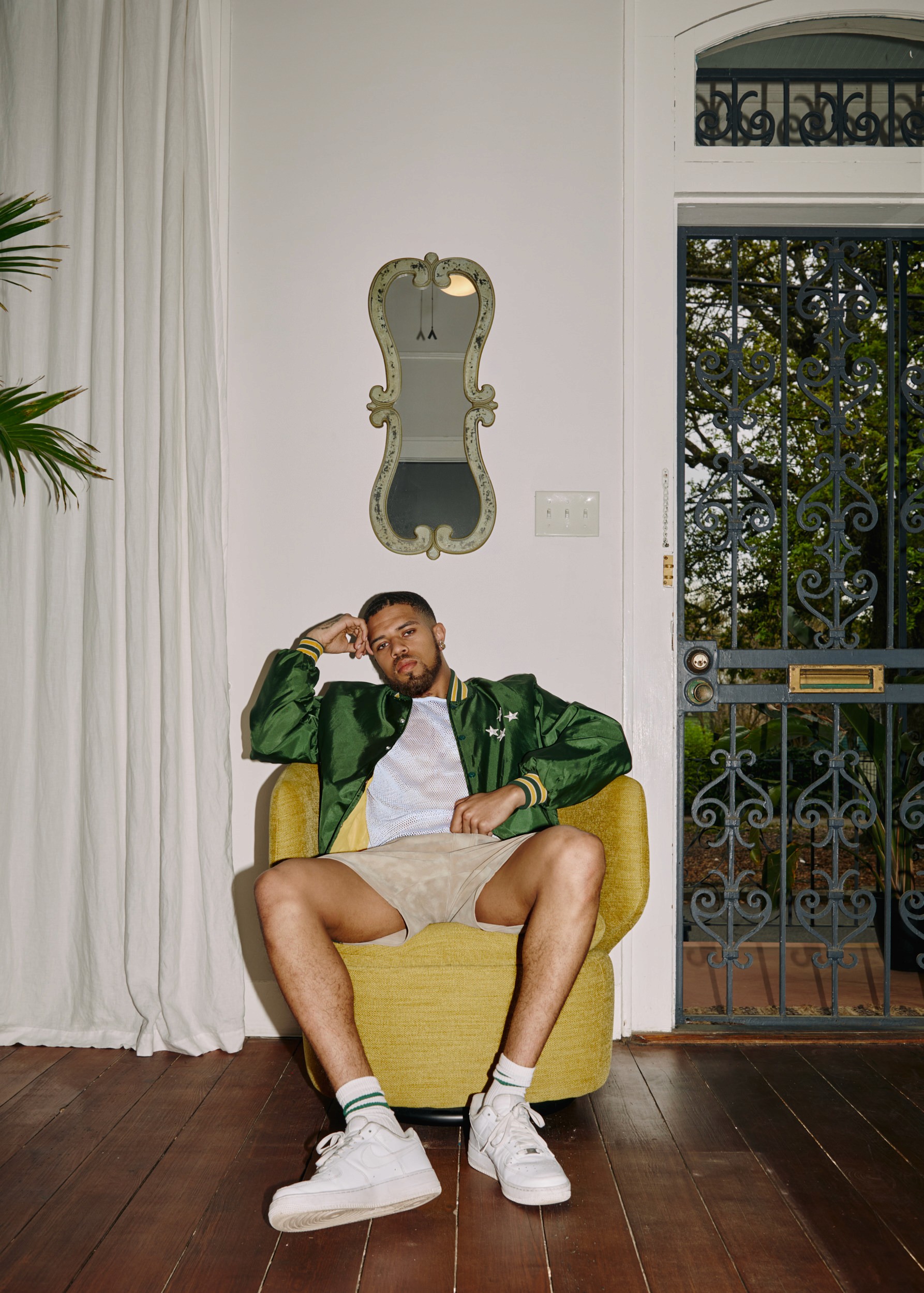 A man in a green bomber jacket sits relaxed in a yellow chair inside a New Orleans home, iron gate visible through the door behind him.