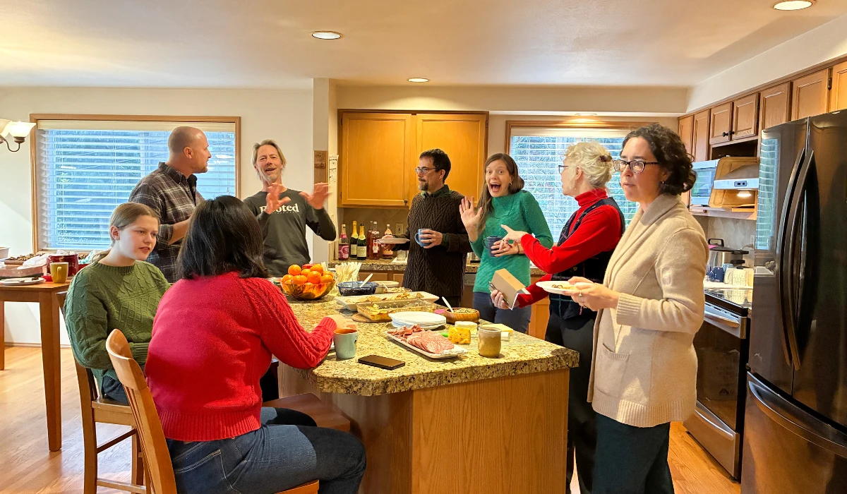 Community members standing and sitting around a kitchen island, talking and laughing together during a shared gathering.