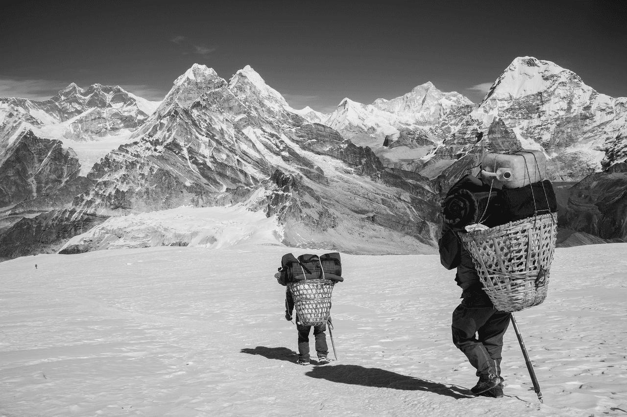 Black and white photograph of Sherpa guides carrying traditional dokos through a Himalayan mountain pass, representing the spirit and heritage behind Café Sherpa's in Brookfield, WI. A café built from years of travel across cultures and continents.