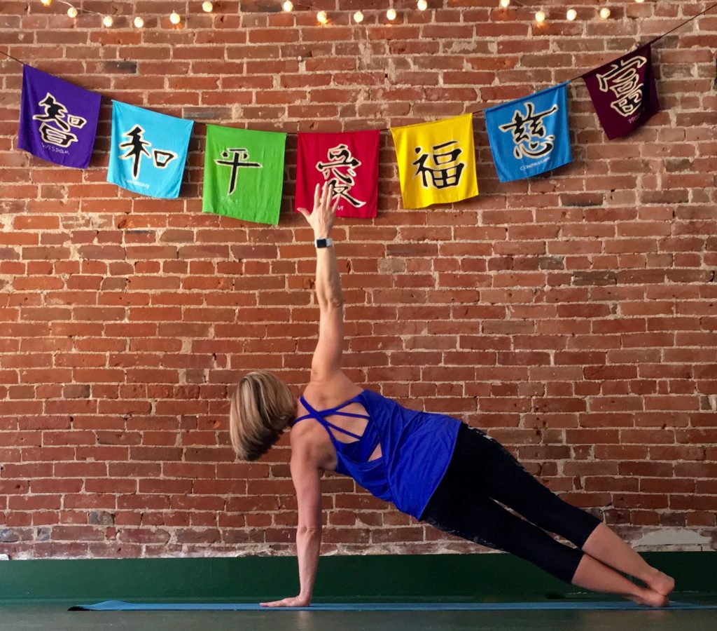 Heather Hausman in athletic wear is performing a forearm plank on a yoga mat indoors, with a background of exposed brick walls.