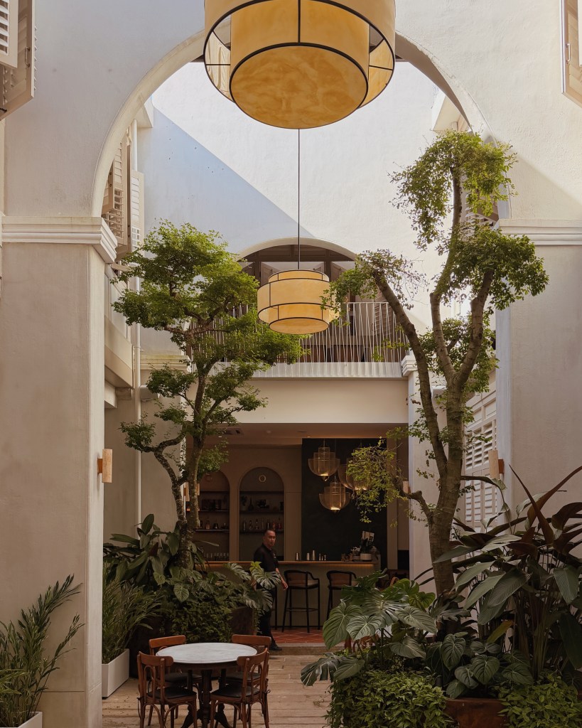 Interior view of a modern courtyard with tall plants, pendant lighting, and a bar area visible in the background.
