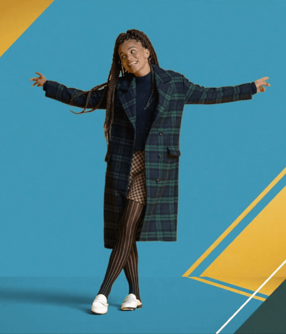 Woman in plaid coat and patterned skirt standing against bright blue and gold geometric backdrop for Nordstrom Rack Fashion at a Fraction campaign.