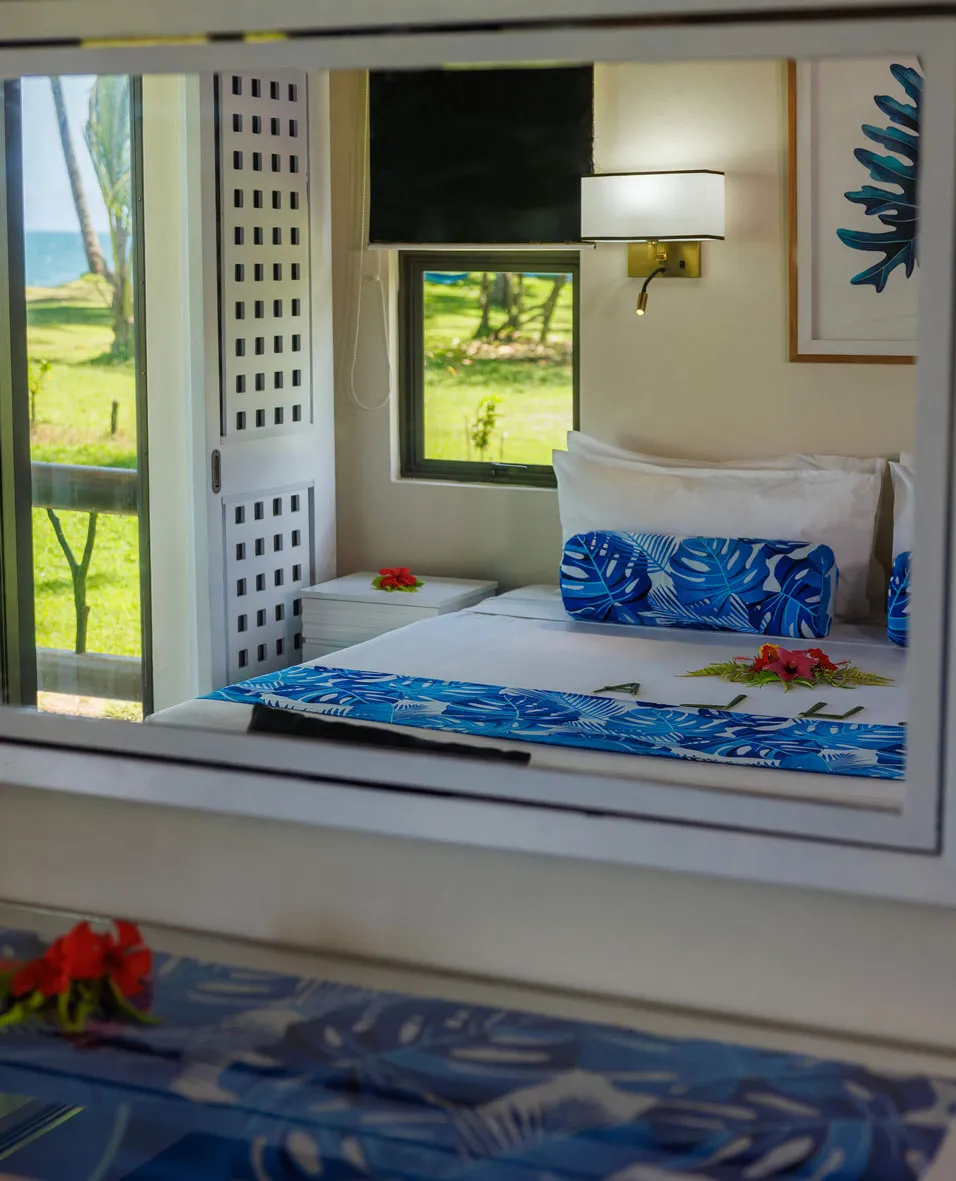 Beachfront bedroom with blue tropical linens and ocean view, reflected in mirror, Uprising Beach Resort.