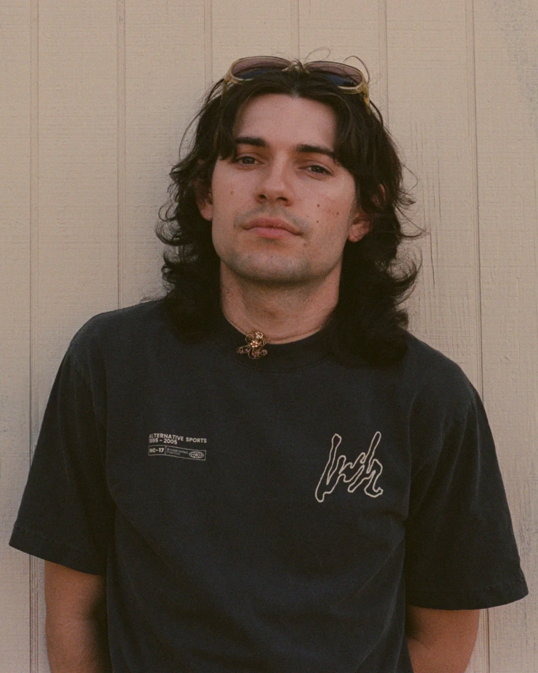 Model standing against a beige paneled wall wearing the grey Alternative Sports 01 T-Shirt with the LWH mark and advisory-style label printed on the chest.