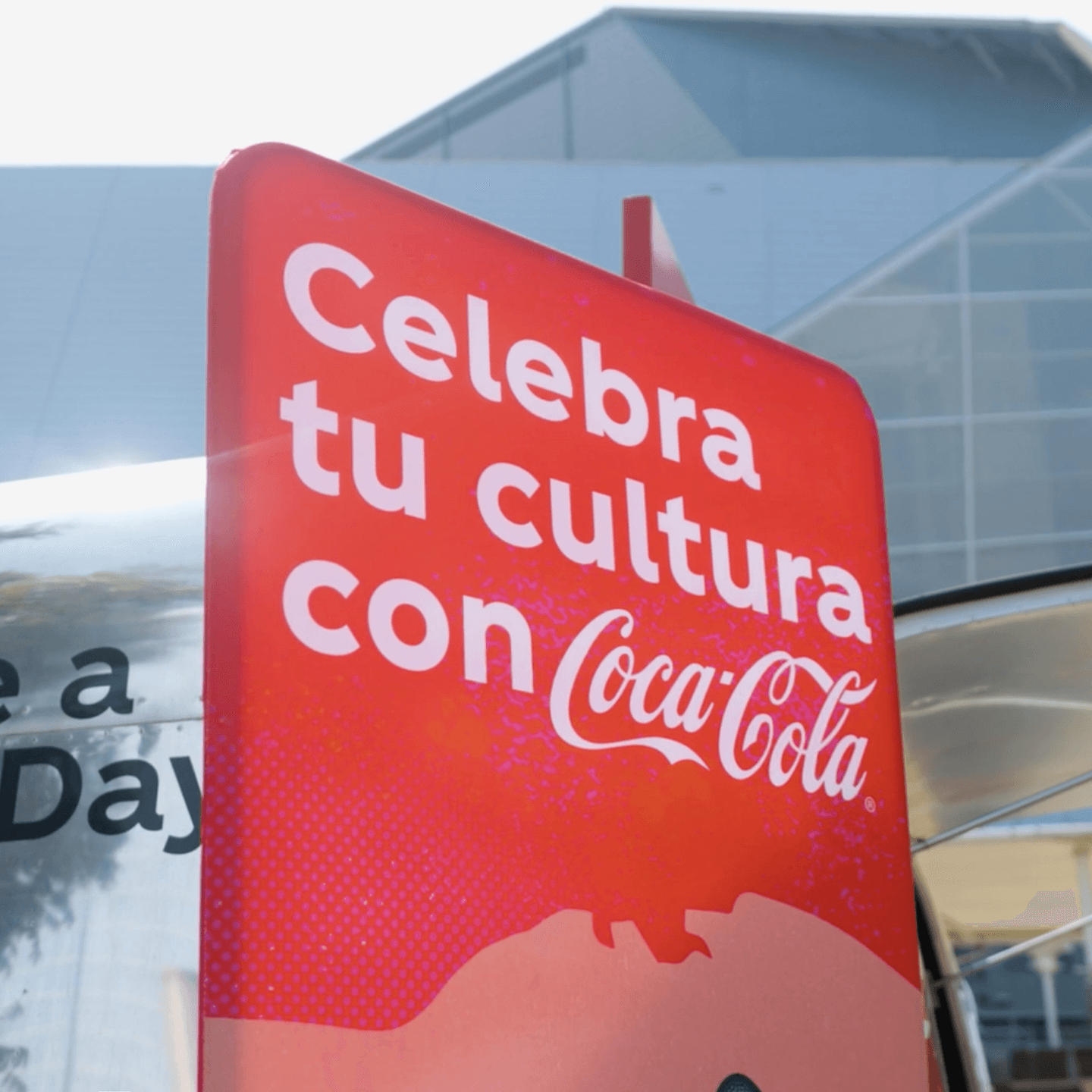 A red Coca-Cola sign reads Celebra tu cultura con Coca-Cola in white text, set against a modern building background.