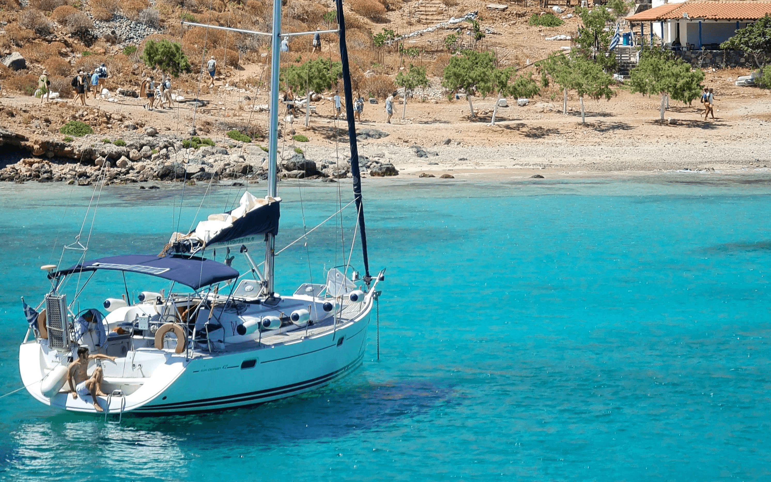 A white sailboat anchored in crystal-clear turquoise water near a rocky coastline.