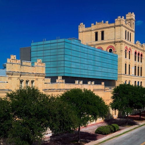 Large building with tan stone brickwork and blue glass modern extension, surrounded by trees under a clear blue sky.