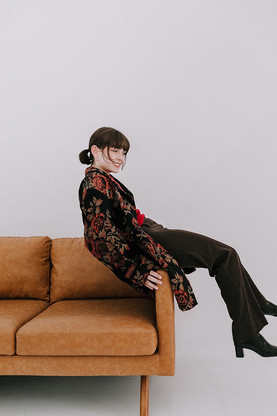 A model sitting on the back of a tan couch, wearing a floral-patterned jacket, part of a 90s-inspired photoshoot at Revelator Studio, a natural light studio in Shreveport.