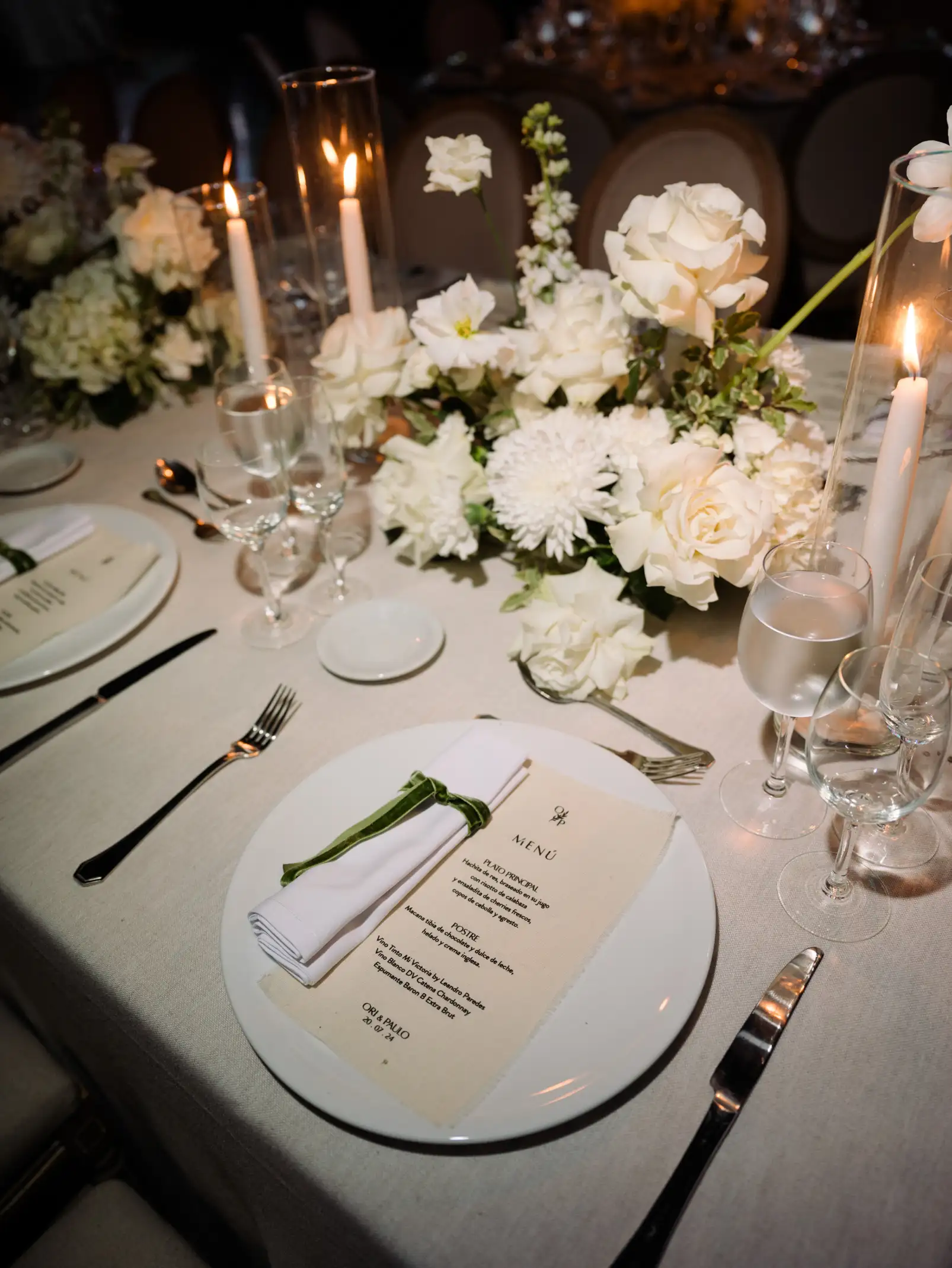 Detalle de mesa con menú, servilleta con cinta verde y velas entre flores blancas.