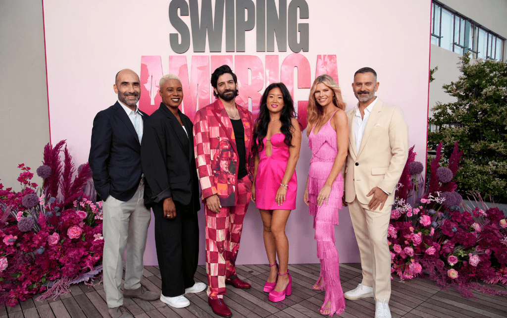 A group of five stylish individuals, the cast of Swiping America, pose together in front of pink flowers and a pink backdrop at an event.