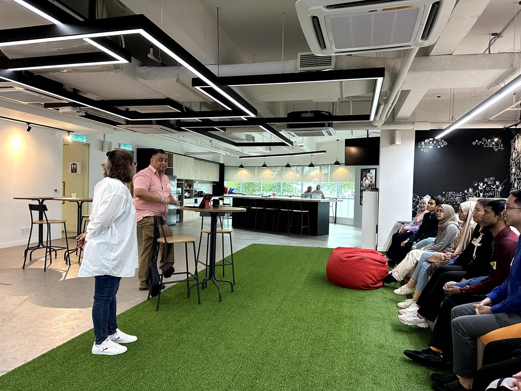 A modern workspace with a grassy area, a speaker presenting to an audience seated on chairs, and a kitchen visible in the background.
