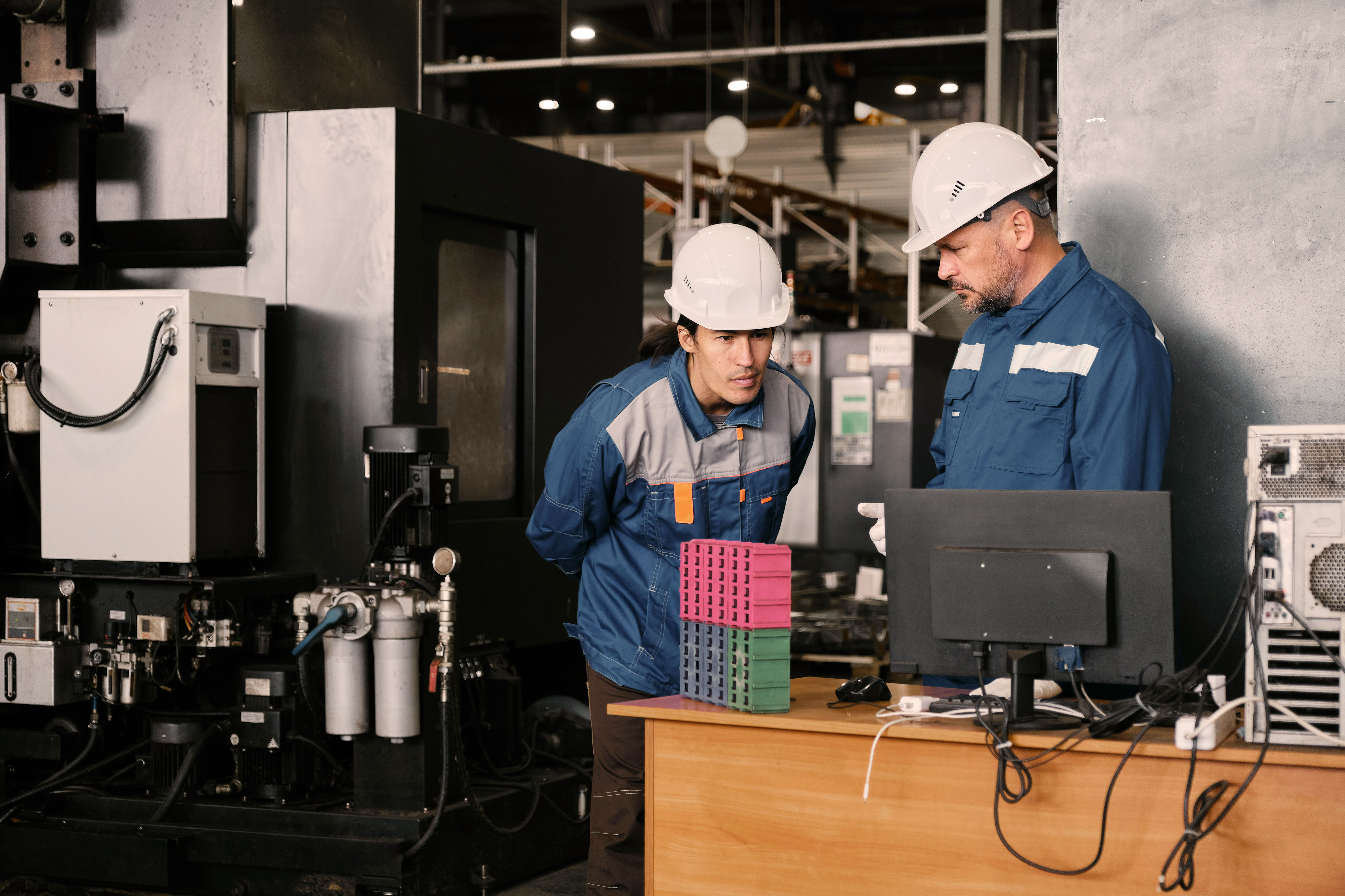 Two workers in hard hats examine a computer screen in an industrial setting, surrounded by machinery.