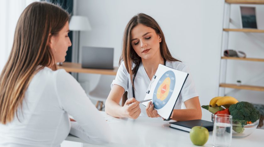 Two women sit at a table discussing a nutritional chart, surrounded by fresh fruits and vegetables, emphasizing the importance of physical health and vitality.
